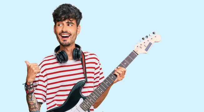 Young hispanic man playing electric guitar pointing thumb up to the side smiling happy with open mouth