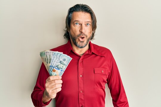 Middle age handsome man holding dollars scared and amazed with open mouth for surprise, disbelief face