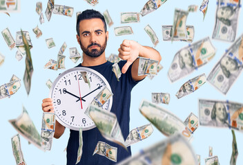 Young hispanic man holding big clock with angry face, negative sign showing dislike with thumbs...