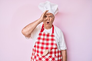 Senior grey-haired man wearing professional baker apron doing ok gesture shocked with surprised face, eye looking through fingers. unbelieving expression.