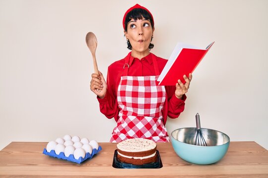 Beautiful brunettte woman cooking cake reading recipes book making fish face with mouth and squinting eyes, crazy and comical.