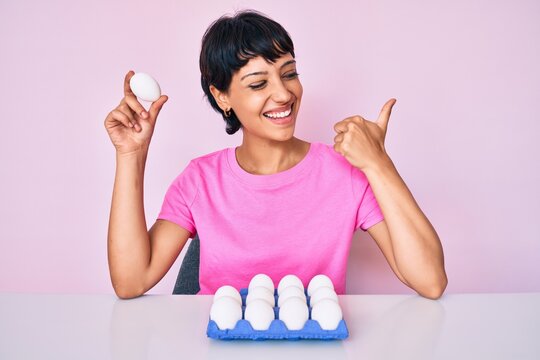 Beautiful brunettte woman showing fresh white eggs pointing thumb up to the side smiling happy with open mouth
