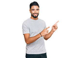 Young man with beard wearing casual grey tshirt smiling and looking at the camera pointing with two hands and fingers to the side.
