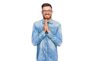 Young redhead man wearing casual denim shirt praying with hands together asking for forgiveness smiling confident.