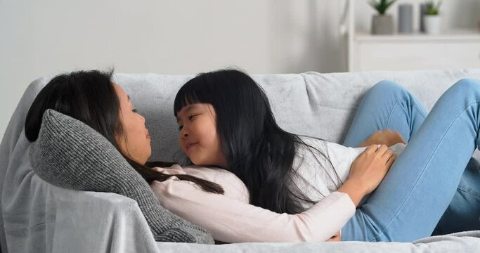 Happy carefree asian korean mother and small child daughter lying on couch sofa laughing having fun smiling together, kid kissing mum on nose enjoying bonding cuddling hugging at home on weekend 