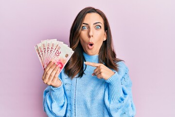 Young brunette woman holding 20 israel shekels banknotes surprised pointing with finger to the side, open mouth amazed expression.