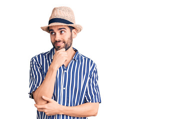 Young handsome man with beard wearing summer hat and striped shirt with hand on chin thinking about question, pensive expression. smiling with thoughtful face. doubt concept.