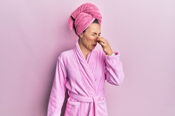 Young blonde woman wearing shower towel cap and bathrobe smelling something stinky and disgusting,...