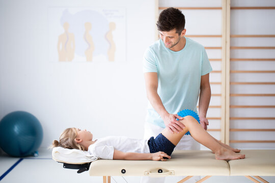 Cute little injure girl on massage table exercising with young male doctor