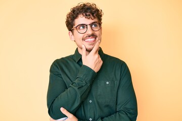 Young caucasian man with curly hair wearing casual clothes and glasses with hand on chin thinking about question, pensive expression. smiling with thoughtful face. doubt concept.