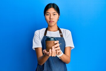 Young chinese woman wearing waiter apron holding cup of coffee relaxed with serious expression on face. simple and natural looking at the camera.