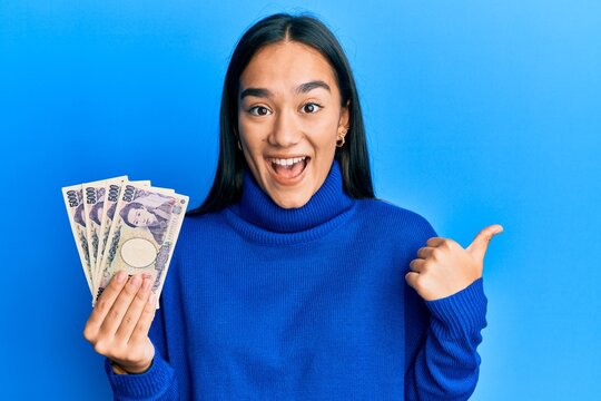 Young asian woman holding 5000 japanese yen banknotes pointing thumb up to the side smiling happy with open mouth