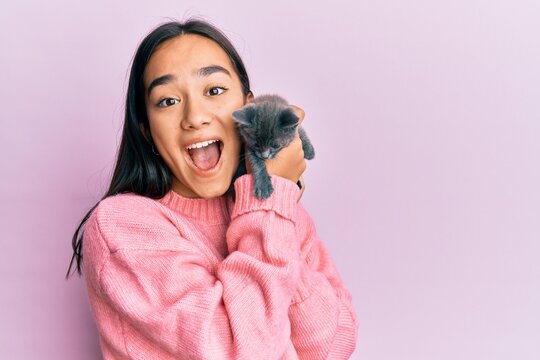Young hispanic girl with surprised face holding cute cat over isolated pink background.