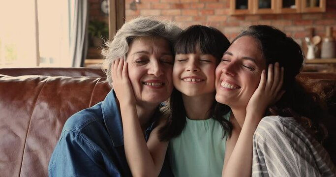 Grandma, mom and daughter of sit on sofa at home smile look at camera. Cute girl touch cheeks of mom and elderly granny enjoy moment of tenderness. Three generations of women family portrait concept