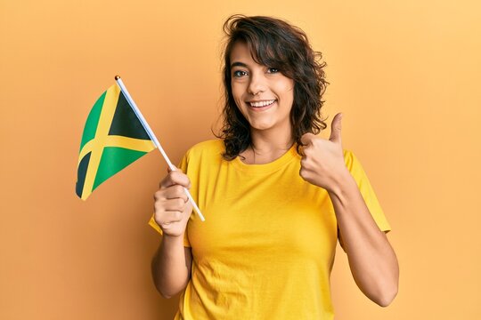 Young hispanic woman holding jamaica flag smiling happy and positive, thumb up doing excellent and approval sign