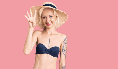 Young blonde woman with tattoo wearing bikini and summer hat showing and pointing up with fingers number four while smiling confident and happy.