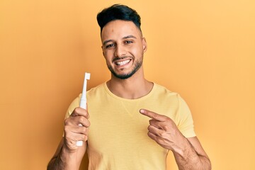 Young arab man holding electric toothbrush smiling happy pointing with hand and finger