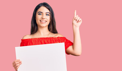Fototapeta premium Brunette teenager girl holding blank empty banner surprised with an idea or question pointing finger with happy face, number one