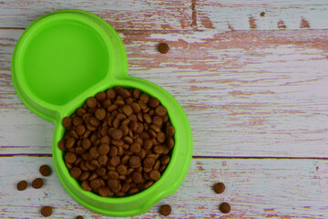 Against a background of gray wooden boards, lies a green plastic plate filled with dry food for feeding cats or dogs. There is a place on the right for an inscription