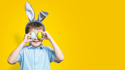 Happy child wearing bunny ears and holding colorful easter eggs in front of his eyes on a yellow background. Happy boy in easter bunny costume with easter eggs