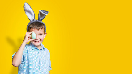 Happy child wearing bunny ears and holding colorful easter egg in front of his eye on a yellow background. Happy boy in easter bunny costume with easter egg