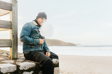 man vaping his electronic cigarette sitting on a wall in front of the beach, fuming and looking at the camera. alternative to tobacco.