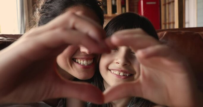 Happy beautiful mother and little 10s adorable daughter faces seen through joined fingers making heart shape, close up. Symbol of love and cherish of mom and kid, donation, sincere feelings concept