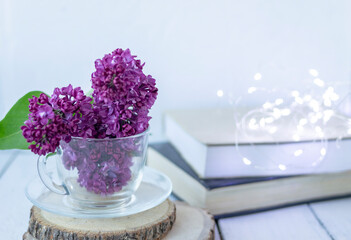 Transparent cup with flowers. Beautiful lilac fills the glass. The spring bouquet is worth no wooden cuts. Macro image of spring lilac purple flowers. Book in the background and lights. Blurred.