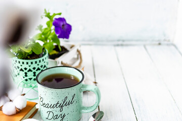Close-up of a cup of tea on a wooden white table with blurred background, front blur. Still life with flower, book, teaspoon, cotton box. Spring breakfast. Copy space.