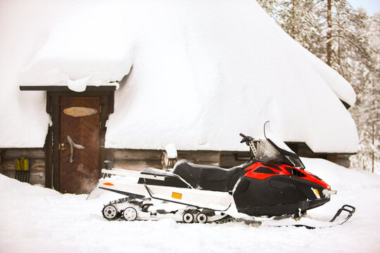Red snowmobile is standing on a snow on the background of a winter house log hut and forest.