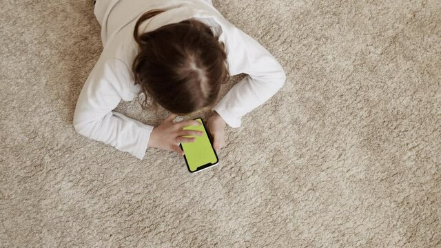 little girl with a phone in her hand, lying on the carpet.Green screen mobile phone. High quality 4k footage