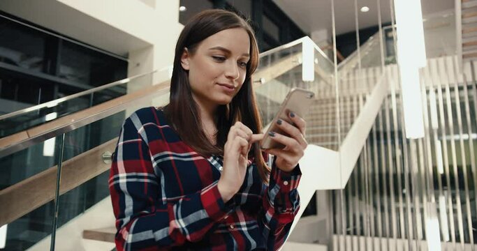 Young beautiful Caucasian female student standing in lobby of library and communicates with friends on social networks using smartphone.