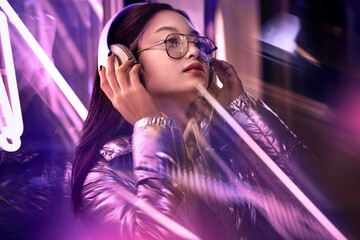 Neon Style. Asian girl in glossy jacket eyeglasses and headphones standing on the street in...