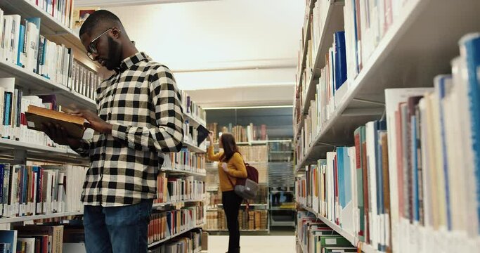 Young smart students stand in modern university library among bookshelves and look for necessary literature to study.
