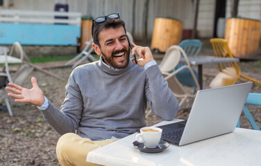 Smiling man working on laptop and talking on smartphone in the coffee bar.
