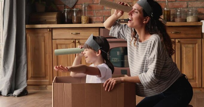 Small girl sit inside of carton box play pirates with mom in kitchen, wear drawn skull on caps, use paper tube like binocular watching into distance pretend be explorers. Playtime, fun at home concept