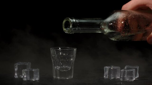 Bartender pouring up shot of vodka with ice cubes from bottle into drinking glass against black background. Barman pour clear transparent alcohol drink vodka tequila in shot-glass. Slow motion footage