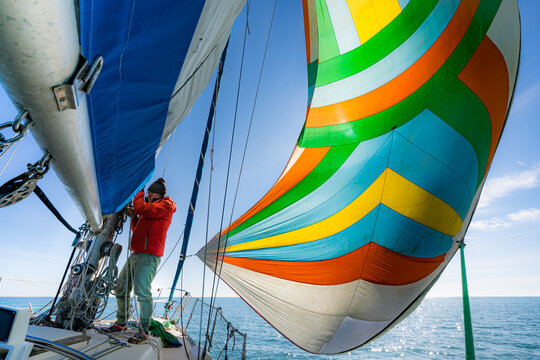 Sur un voilier , un homme en anorak rouge travaille sur le pont entre deux grandes voiles