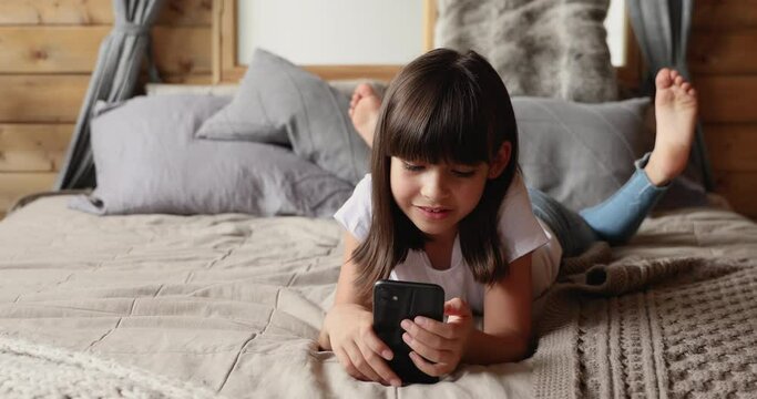 Cute small 10s girl with stylish bob with bangs hairstyle lying on bed holding smartphone device, spend time on internet, watch online channels, gen z and modern tech, mobile application usage concept
