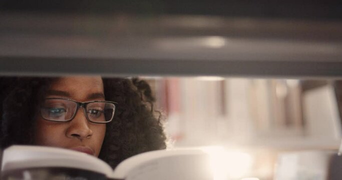 Close up of young beautiful smart african american woman standing in library among shelves and intently reading book.