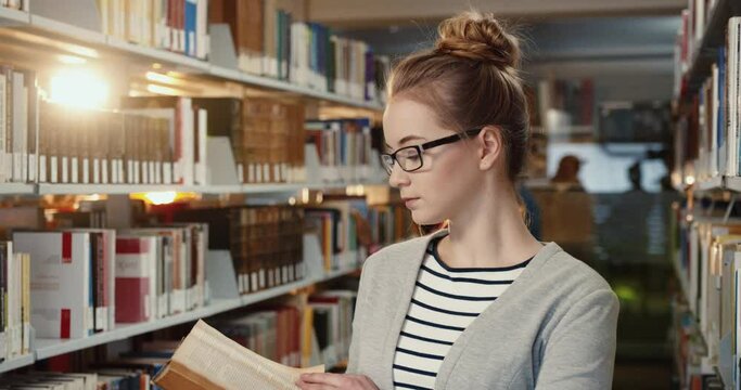Close up of young smart student girl with glasses standing in reading room among shelves looking at camera smiling.