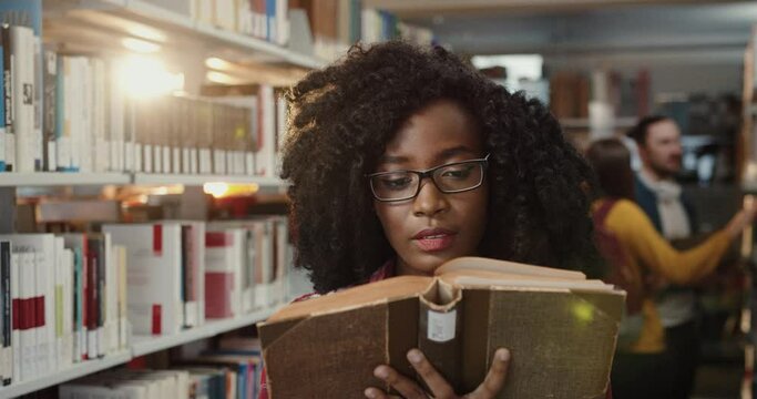Close up of young beautiful african american girl standing in library intently reading book adjusting her glasses and turning the page.