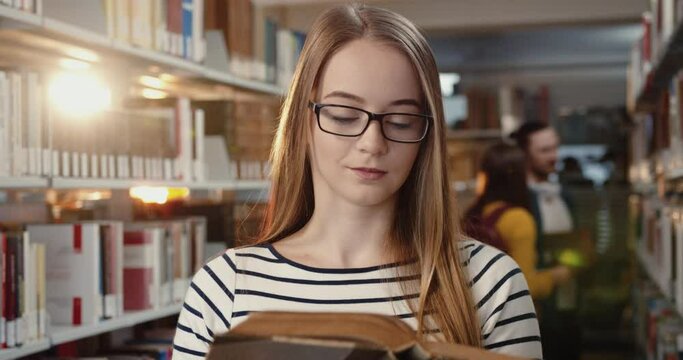 Close up of young beautiful caucasian girl takes necessary book from shelf in library and compares the information with nod.
