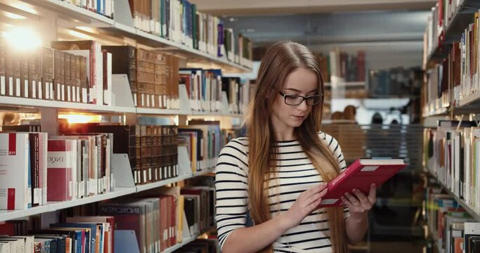 Young smart caucasian blonde girl with glasses stands among bookshelves and chooses literature to read at leisure.