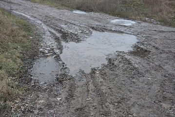 part of a rural road of gray earth with a large pothole and frozen muddy water in the ice