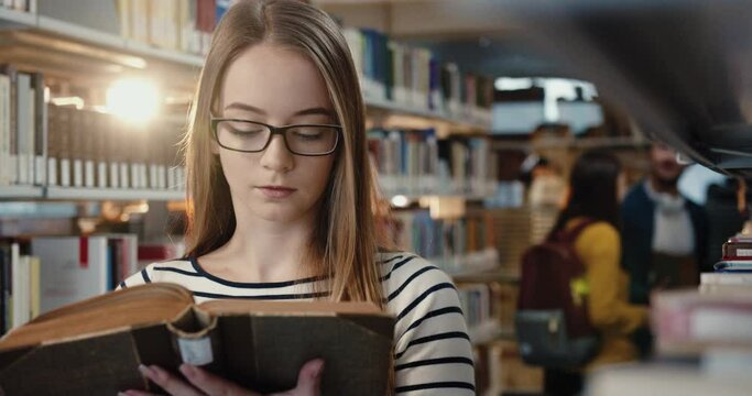 Close up of young smart blonde female student in glasses standing in library and intently studying book.