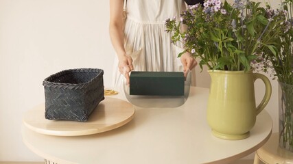 Mujer sosteniendo una caja de cartón. Adornos sobre la mesa. Canasta y jarrón verde con flores