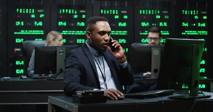 African American IT worker sitting in server room behind the workplace talking on smartphone solving work issues working with database on computer.