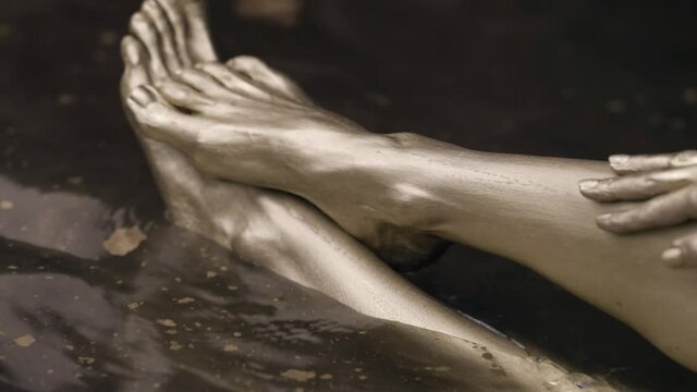 Close-up of slender female legs, submerged in water, covered with gold paint. She touches them with her hands.