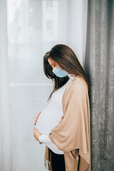 Fototapeta premium Young pregnant woman is standing by window with face protective mask. She is worried about childbirth because od Covid-19 pandemic.